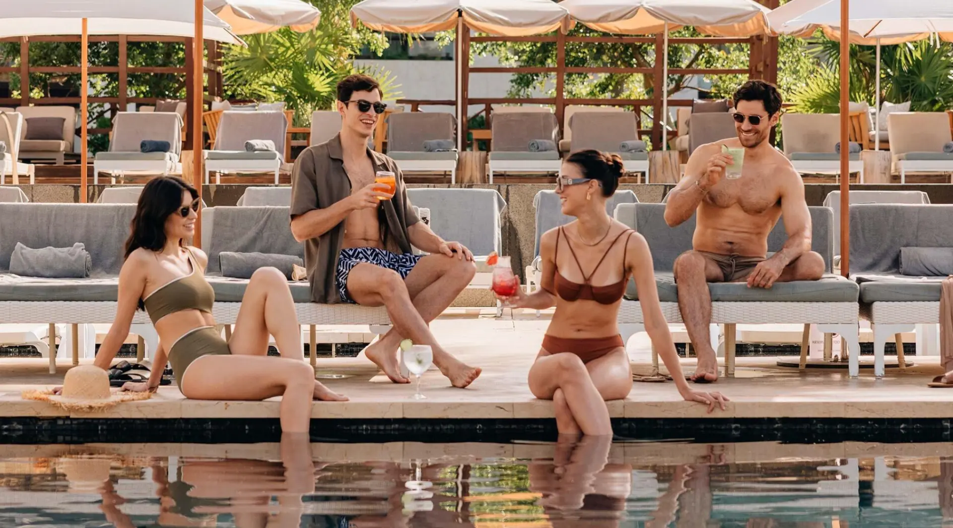 Four stylish adult friends enjoying drinks beside a resort pool, with a man in an open shirt and a shirtless man